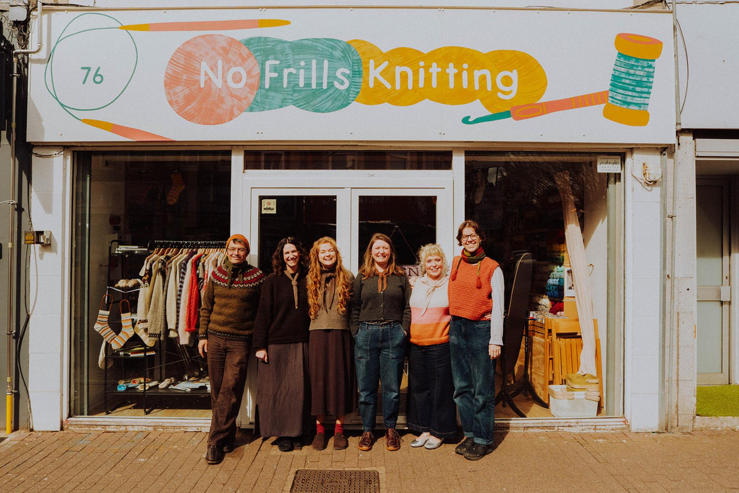 a group shot of the no frills knitting team outside the front of the shop. They are lined up, wearing their handmade knits and all smiling at the camera